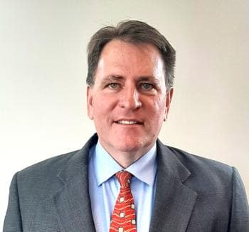 Middle aged Caucasian male with a slight smile wearing a reddish tie, light blue button down shirt and dark patterned suit coat.